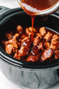 A black slow cooker on a white marble cooktop, as the freshly prepared barbecue sauce is being poured over the chicken. The thick, rich sauce cascades over the chicken, coating each piece evenly for a flavorful slow-cooked dish.