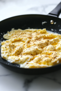 The smooth egg and cottage cheese mixture being poured evenly into the black non-stick skillet over the white marble cooktop, spreading out to form the base of the flatbread.