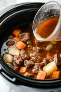 A close-up shot of a measuring cup pouring rich beef broth and Worcestershire sauce into the black slow cooker on the white marble cooktop. The golden-brown liquid cascades over the beef and vegetables, soaking them in a deep, savory flavor.