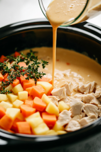 A close-up shot of a glass measuring cup on the white marble cooktop, pouring thick, golden condensed cream of chicken soup over the layered ingredients in the black slow cooker. The creamy mixture evenly coats the chicken and vegetables, adding richness to the dish.