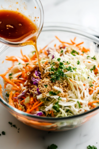 A close-up shot of a small glass bowl pouring the freshly whisked Asian dressing over the shredded cabbage, carrots, and crushed ramen noodles in the large glass mixing bowl on the white marble cooktop, evenly coating the salad ingredients.