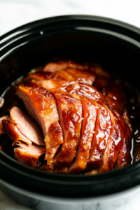 A close-up shot of the brown sugar maple glaze being poured evenly over the spiral-cut ham inside the black crockpot on the white marble cooktop. The syrupy mixture seeps into the slices, creating a glossy sheen and infusing the ham with rich, sweet flavors.