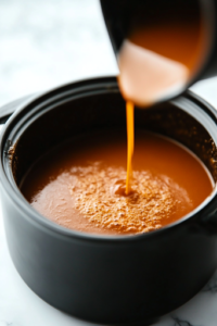 A black crockpot on a white marble cooktop as the cornstarch slurry is being poured into the simmering sauce. The mixture blends in smoothly, thickening the sauce into a rich, glossy coating for the chicken.