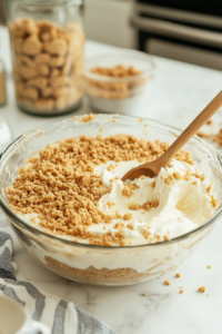A glass mixing bowl filled with crushed graham crackers, sugar, and melted butter being mixed together with a spatula on a white marble countertop.