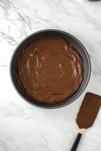 An 8x8 baking pan on a white marble cooktop, with smooth chocolate cake batter spread evenly across the bottom. A spatula rests nearby, coated in chocolate.