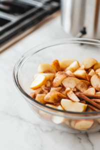A glass mixing bowl on a white marble cooktop filled with sliced apples, sugar, and cinnamon. A wooden spoon rests beside the bowl as the apples are tossed with the spices.
