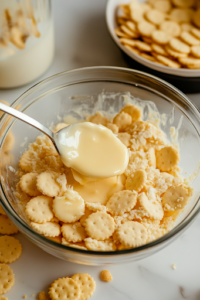A separate glass bowl on a white marble cooktop, filled with crushed buttery round crackers. Melted butter is being poured over the top, and a spoon is stirring the mixture, coating each crumb with rich, golden flavor.