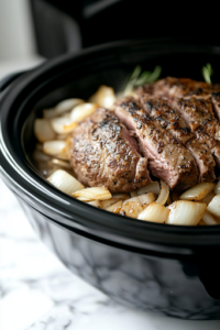 A close-up of a black slow cooker on a white marble cooktop, as a layer of freshly sliced onions is spread at the bottom. The seared pork loin rests on top, ready to soak in the flavors during the slow cooking process.
