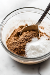A small bowl with white sugar, cornstarch, ground ginger, and cinnamon on a wooden countertop. A spoon is stirring the dry ingredients, evenly combining them for the apple filling.
