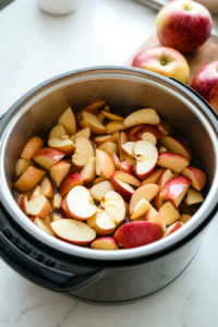 Freshly peeled, cored, and chopped apples in a slow cooker on a wooden countertop. A hand is sprinkling the cornstarch mixture over the apples, ensuring they are evenly coated. Lemon juice is drizzled over the top.