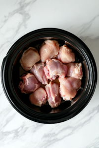 A black crockpot on a white marble cooktop, filled with raw boneless, skinless chicken thighs neatly arranged inside. The chicken has a fresh, light pink hue and is ready for slow cooking.