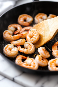 A close-up shot of raw, peeled, and deveined shrimp in a black skillet over a white marble cooktop. The shrimp are beginning to cook, turning slightly opaque, while a wooden spatula stirs them evenly in the sizzling butter.