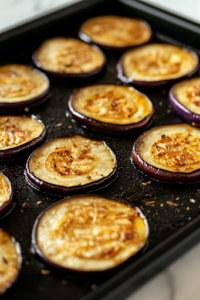 A black baking sheet lined with evenly spaced eggplant slices brushed with olive oil. The edges of the slices are turning golden brown after roasting at 400°F, resting on a white marble cooktop.