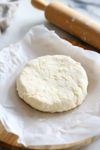Dough placed between two sheets of parchment paper on a white marble cooktop. A rolling pin gently presses down, flattening it into a circular shape.