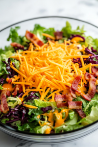 A close-up of the freshly mixed salad, topped with shredded cheddar cheese and crispy bacon pieces. The melted cheese and crunchy bacon contrast beautifully with the creamy salad base.