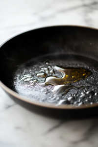 Close-up shot of a black skillet on the white marble cooktop, with olive oil and butter sizzling. Fresh broccoli florets are being sautéed until crisp-tender, with a golden-brown crust forming on some of the edges.