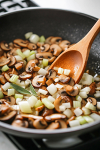 A skillet sizzling with diced onions, celery, and sliced mushrooms being sautéed in olive oil. The vegetables are softened and fragrant, with minced garlic and fresh sage being stirred in for extra flavor.