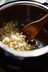 A close-up shot of an Instant Pot on a white marble cooktop, set to 'Sauté' mode. Minced garlic sizzles inside, releasing a rich aroma, while a wooden spoon stirs gently to evenly distribute the heat.