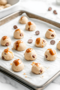 A close-up shot of a parchment-lined baking sheet on the white marble cooktop, featuring evenly spaced cookie dough scoops placed 2 inches apart. The dough mounds have a slightly textured surface, with visible bits of caramel and chocolate chips.