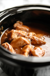A black crockpot on a white marble cooktop as the rich, flavorful honey garlic sauce is being poured over the raw chicken. The sauce coats the meat evenly, pooling slightly at the bottom, ensuring every piece absorbs the bold flavors.