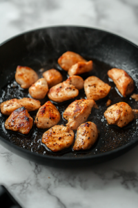 A black skillet on a white marble cooktop with golden-brown chicken pieces sizzling in hot olive oil. Light steam rises as the chicken sears, creating a crispy, flavorful outer layer.