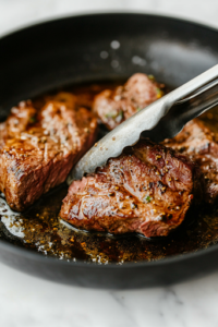 A close-up of a black skillet placed on a white marble cooktop, where the seasoned chuck roast is being seared in sizzling olive oil. The surface of the meat develops a deep golden-brown crust as a pair of tongs holds it in place.