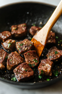 A close-up shot of the black skillet on the white marble cooktop, where the steak bites are searing to a deep golden-brown crust. A pair of tongs is flipping a piece, revealing a perfectly caramelized surface that glistens with rendered juices.