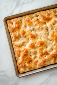 A baking sheet sits on a white marble cooktop, holding focaccia dough sprinkled with flaky sea salt and freshly chopped rosemary. The dough is well-seasoned and ready to go into the oven.