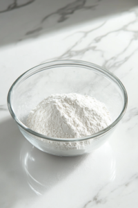 A glass mixing bowl on a white marble cooktop containing a blend of all-purpose flour, paprika, salt, and black pepper. A hand is seen whisking the ingredients together, ensuring an even coating mixture for the fried chicken.
