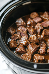 A close-up of a black slow cooker on a white marble cooktop, as a hand evenly sprinkles a packet of au jus gravy mix and onion soup mix over the steak bites. The seasoning coats the meat, ensuring deep, rich flavors during the cooking process.