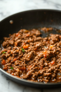 A black skillet on a white marble cooktop with browned ground beef as taco seasoning and water are being stirred in. The mixture simmers, turning into a rich, flavorful filling with a slightly thickened consistency.