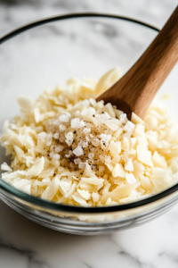 A close-up shot of minced garlic, dried oregano, sea salt, and freshly ground black pepper being sprinkled into the glass mixing bowl on the white marble cooktop. The aromatic seasonings enhance the vinaigrette with bold Mediterranean flavors.