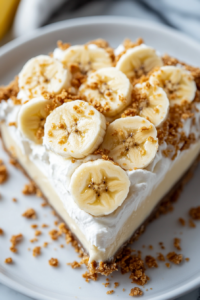 A beautifully sliced piece of banana cream pie on a plate, topped with coconut whipped cream and fresh banana slices. A fork rests beside it, ready for a bite.