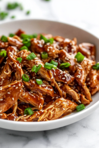 A white plate on a white marble cooktop, filled with shredded honey garlic chicken served over cauliflower rice. The dish is garnished with toasted sesame seeds and freshly chopped green onions, adding a final touch of flavor and color, ready to be enjoyed.