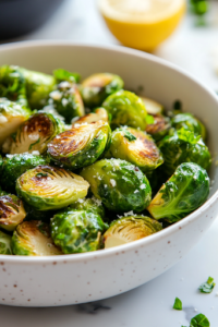A close-up shot of a white serving bowl on a white marble countertop, filled with bright green, glistening Brussels sprouts. A drizzle of olive oil, fresh parmesan, and a squeeze of lemon complete the dish, ready to be enjoyed.
