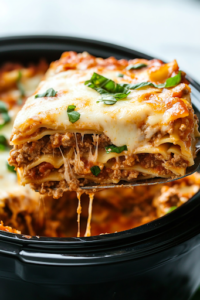 A close-up shot of a slice of slow-cooked lasagna being lifted from the black slow cooker with a spatula over the white marble cooktop. The layers of noodles, melted cheese, and savory meat sauce hold together beautifully, with fresh basil sprinkled on top for a perfect finishing touch.