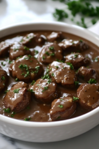 A close-up of a rustic serving dish on a white marble cooktop, filled with juicy steak bites coated in a savory, glossy gravy. A final sprinkle of freshly chopped parsley adds a vibrant touch, enhancing the dish before serving.