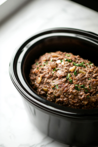 A close-up of a black slow cooker on a white marble cooktop, as a pair of hands carefully shape the seasoned meat mixture into a firm, compact loaf and place it inside. The loaf sits snugly in the slow cooker, ready for gentle, even cooking.