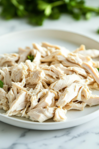 A close-up shot of a white plate on the white marble cooktop, with two forks pulling apart the tender, cooked chicken into bite-sized shreds. The juicy and well-seasoned chicken is ready to be returned to the broth for a hearty soup.