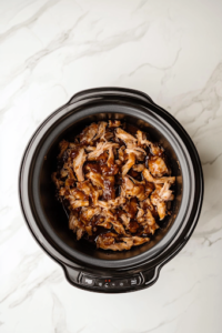 A close-up of a black slow cooker on the white marble cooktop, as a pair of forks shreds the tender, fall-apart pork. The juicy meat glistens in the flavorful BBQ sauce, soaking up every bit of smoky-sweet goodness.