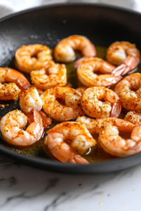 A black skillet on a white marble cooktop with shrimp sizzling as they turn pink. The Cajun seasoning forms a flavorful, slightly charred crust on the shrimp. Tongs hover over the skillet, ready to flip the shrimp.