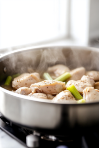 A large stainless steel pot on a white marble cooktop, filled with bone-in chicken breasts, celery stalks, salt, and pepper. The pot is gently simmering, with steam rising as the chicken cooks until tender, infusing the broth with rich flavors.