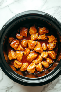 A black crockpot on a white marble cooktop with orange chicken simmering in a thickened sauce after three hours of slow cooking. The sauce glistens as it coats the tender chicken pieces, intensifying the rich, tangy flavor.