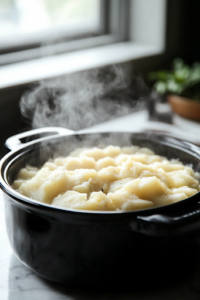 A black crockpot on a white marble cooktop with the potato mixture simmering gently after six hours of slow cooking. Steam rises as the potatoes soften and blend into a creamy consistency, preparing the soup for the next ingredients.