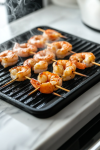A close-up shot of a preheated black grill pan on a white marble cooktop, with marinated shrimp threaded onto metal skewers. The shrimp are arranged neatly, with wisps of steam rising from the hot grill, indicating the cooking process is about to begin.