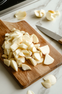 A close-up shot of freshly peeled, cored, and thinly sliced apples neatly arranged on a wooden cutting board over a white marble cooktop. A sharp paring knife rests nearby, with scattered apple peels adding a rustic touch.