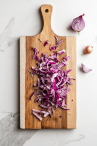 A close-up shot of a sharp knife slicing a red onion thinly on a wooden cutting board over the white marble cooktop, with its vibrant purple layers neatly stacked and slightly curled at the edges, ready for the salad.