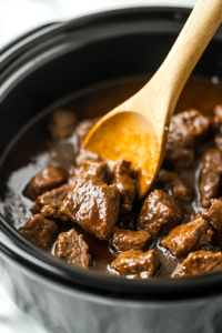 A close-up of a black slow cooker on a white marble cooktop, as the lid is lifted, revealing tender, slow-cooked steak bites glistening in a rich, flavorful gravy. A wooden spoon stirs gently, ensuring the sauce evenly coats each piece