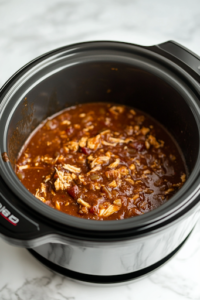 A black slow cooker on a white marble cooktop, covered and cooking on low heat. Steam slightly escapes from the edges, as the chicken slowly simmers in the deep, smoky barbecue sauce, infusing it with bold flavors.