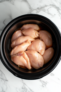 A black slow cooker on a white marble cooktop, with raw boneless, skinless chicken breasts neatly arranged inside. The chicken is placed evenly at the bottom, ready to be transformed into a flavorful and hearty white chicken chili.